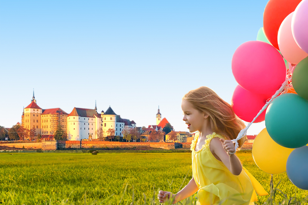 Kind rennt mit bunten Luftballons über Elbwiese mit Schlosshartenfels im Hintergrund