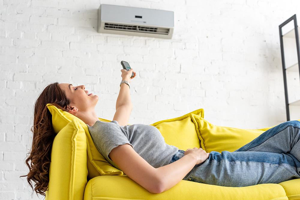 Frau liegt auf einem gelben Sofa und bedient eine Klimaanlage über ihr