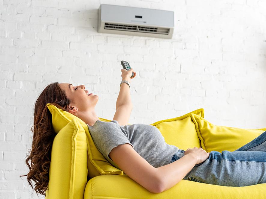 Frau liegt auf einem gelben Sofa und bedient eine Klimaanlage über ihr