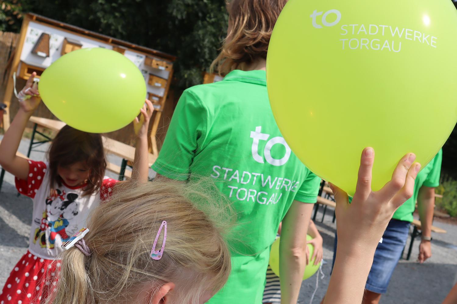 Stadtwerke Torgau Mitarbeiterin mit grünem T-Shirt umringt von Kindern mit grünen Luftballons mit Stadtwerke Torgau Logo