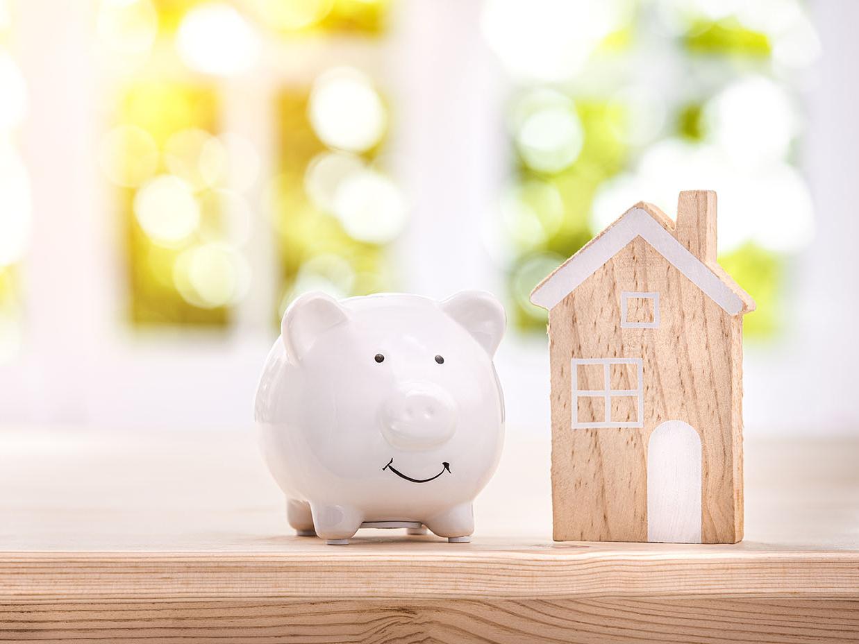 weißes Sparschwein mit Holzhaus steht auf Holztisch vor hellem Fenster